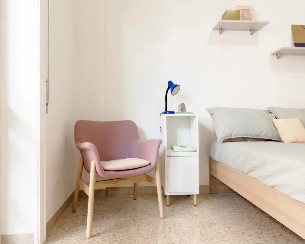 Bright, minimal bedroom corner with a comfortable bed, soft grey pillows and a pastel armchair beside a small white bedside shelf and lamp.