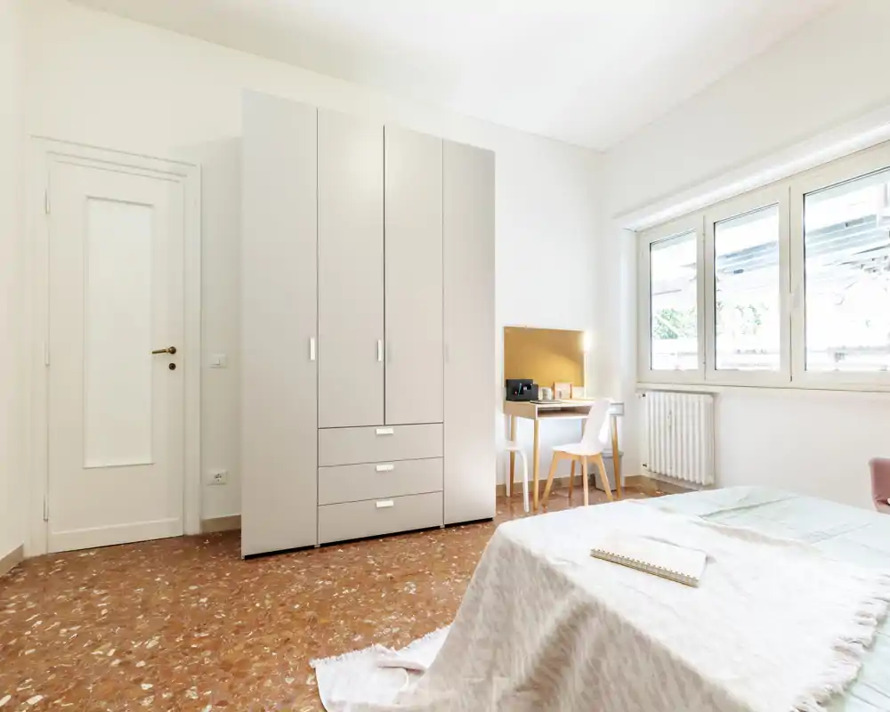 Dormitorio luminoso y minimalista con gran armario, escritorio junto a la ventana y cama bien hecha con textiles suaves — ideal para una estancia tranquila y luminosa.