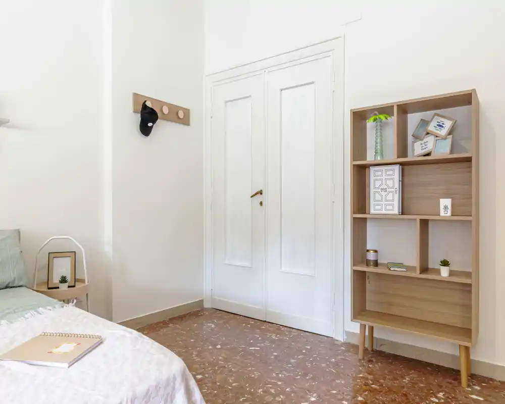 Dormitorio luminoso y minimalista con la cama parcialmente visible, mesita de noche y estantería de madera que ofrecen almacenamiento moderno y sencillo.