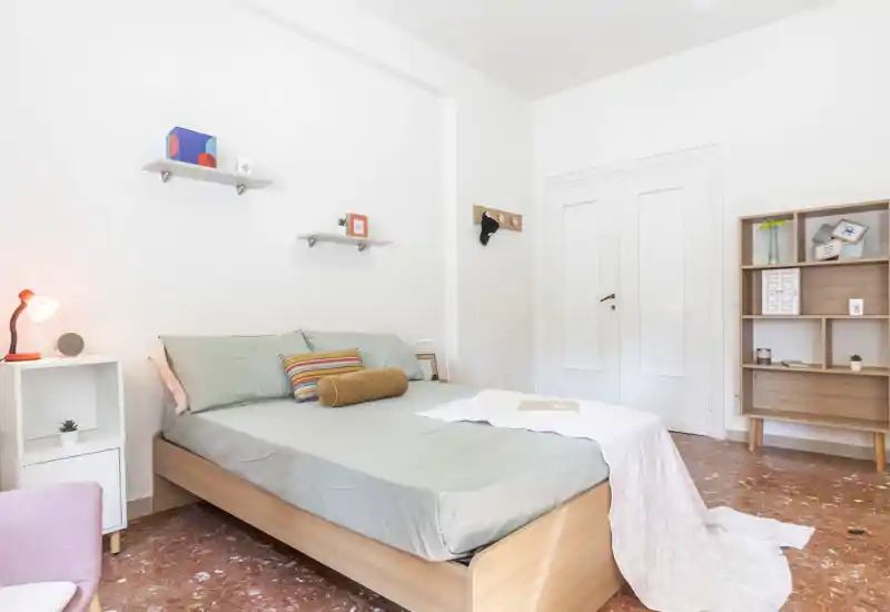 Dormitorio moderno y luminoso con cama doble bien vestida, textiles suaves y estanterías minimalistas: un espacio acogedor y ventilado ideal para alquileres de corta duración.