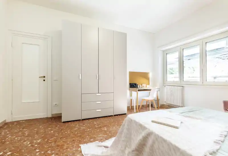 Dormitorio luminoso y minimalista con gran armario, escritorio junto a la ventana y cama bien hecha con textiles suaves — ideal para una estancia tranquila y luminosa.