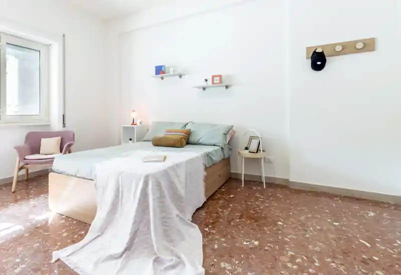 Dormitorio luminoso y ordenado con cama doble de tamaño medio, ropa de cama en tonos pastel, un sillón junto a la ventana y estanterías minimalistas — perfecto para una estancia tranquila y moderna.