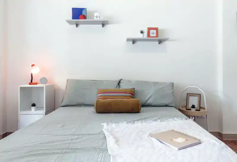 Dormitorio luminoso y minimalista con una cama grande centrada, ropa de cama gris, cojines decorativos y luz cálida de mesita — un espacio para descansar.