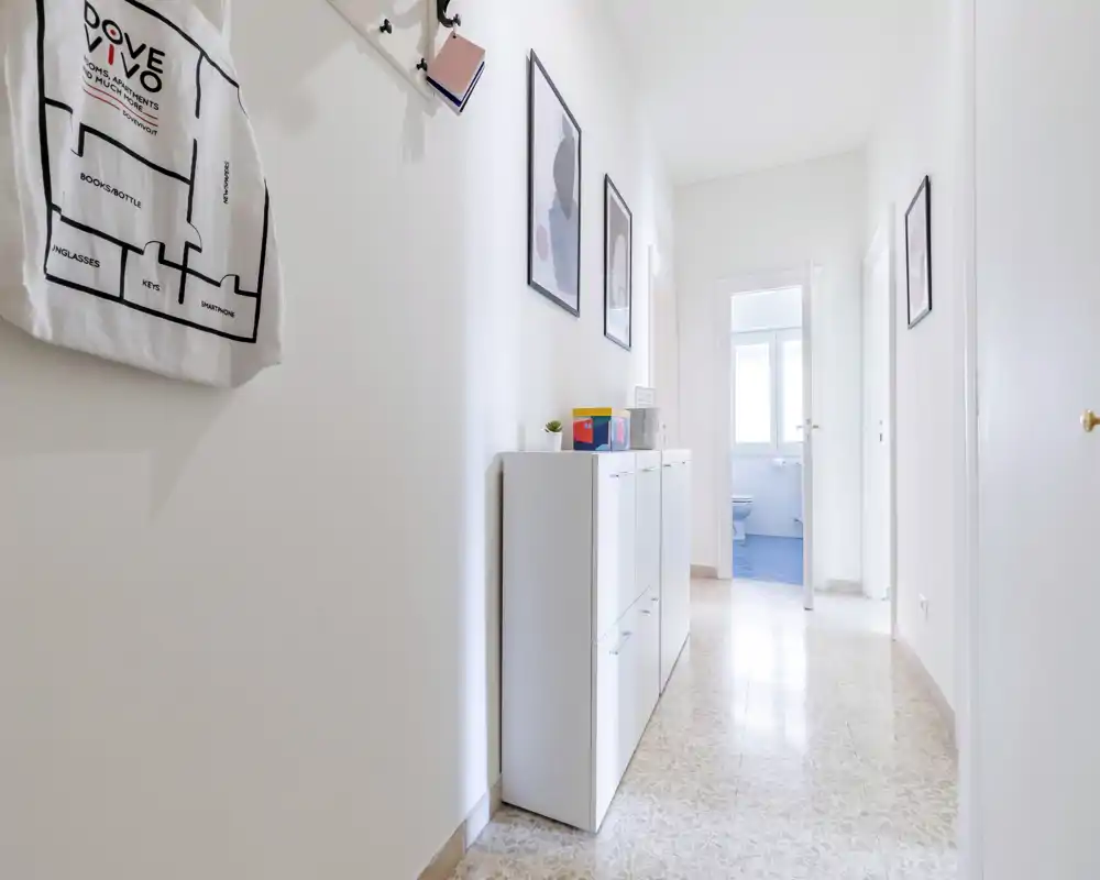 Vestíbulo luminoso y minimalista con paredes blancas, un armario zapatero estrecho y cuadros que conducen a otras estancias. El suelo pulido y la luz natural hacen la entrada acogedora.