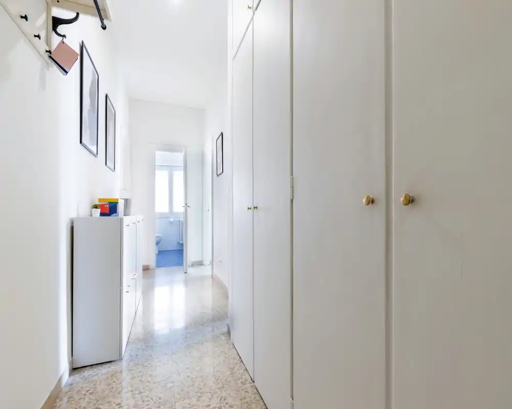 Pasillo de entrada luminoso y limpio con armarios empotrados y zapatero, que conduce al baño al fondo. Decoración neutra y suelo de terrazo pulido que aportan sensación de amplitud.