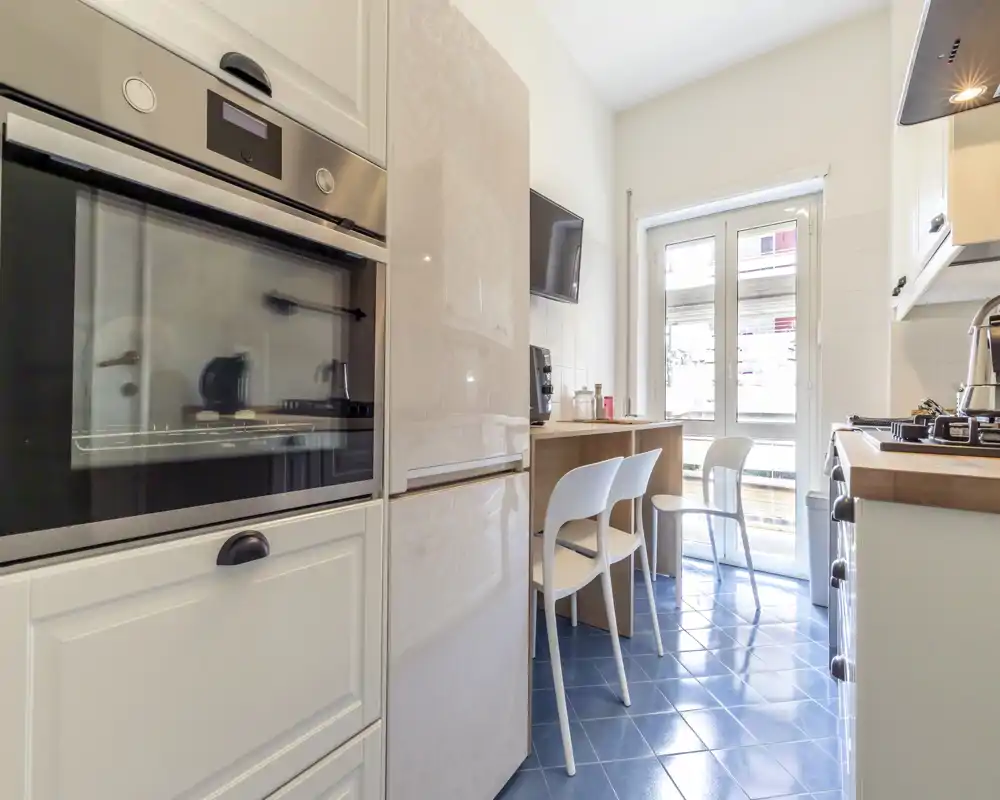 Cocina moderna y luminosa con horno empotrado, frigorífico, pequeño mostrador de madera y sillas junto a una gran ventana que aporta luz natural.
