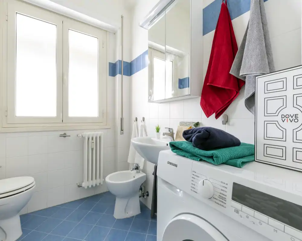 Baño luminoso y moderno con lavadora, bidé, lavabo y armario con espejo; suelo azul y paredes blancas que dan una sensación fresca y limpia.