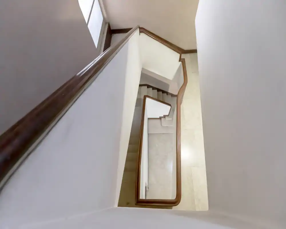 Vista desde arriba de una escalera interior limpia con pasamanos de madera y peldaños de piedra clara; paredes neutras y luz natural de una ventana.