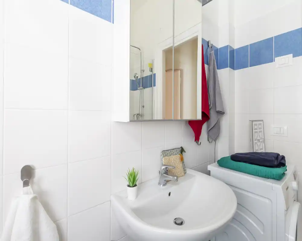Baño moderno y luminoso con azulejos blancos, armario espejo, lavabo y espacio con lavadora/secadora apilada y toallas dobladas.