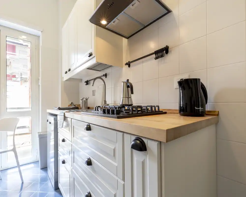 Cocina compacta y luminosa con armarios blancos estilo shaker, encimera de madera, placa de gas y hervidor — práctica y bien presentada para un piso pequeño.