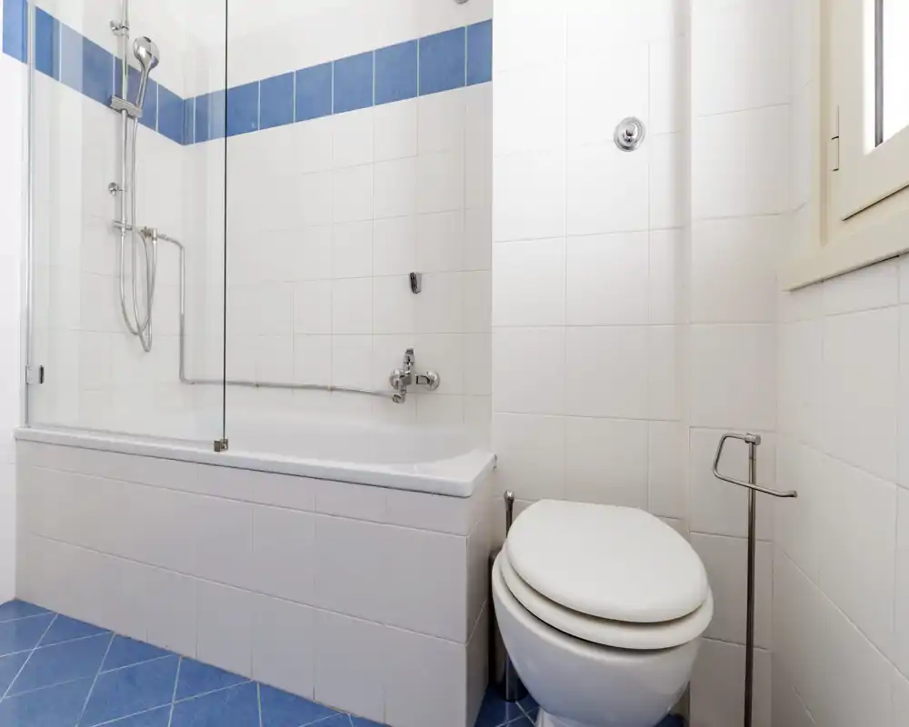 Baño limpio y luminoso con bañera y ducha combinadas, paredes con azulejos blancos y suelo azul, además de un inodoro moderno.