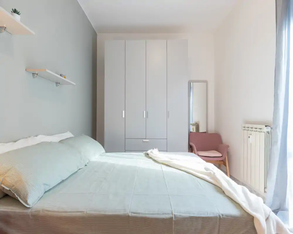Dormitorio doble luminoso y minimalista con cama grande, textiles suaves y armario de tres puertas. La luz natural inunda la habitación, creando una atmósfera tranquila y aireada perfecta para anuncios.