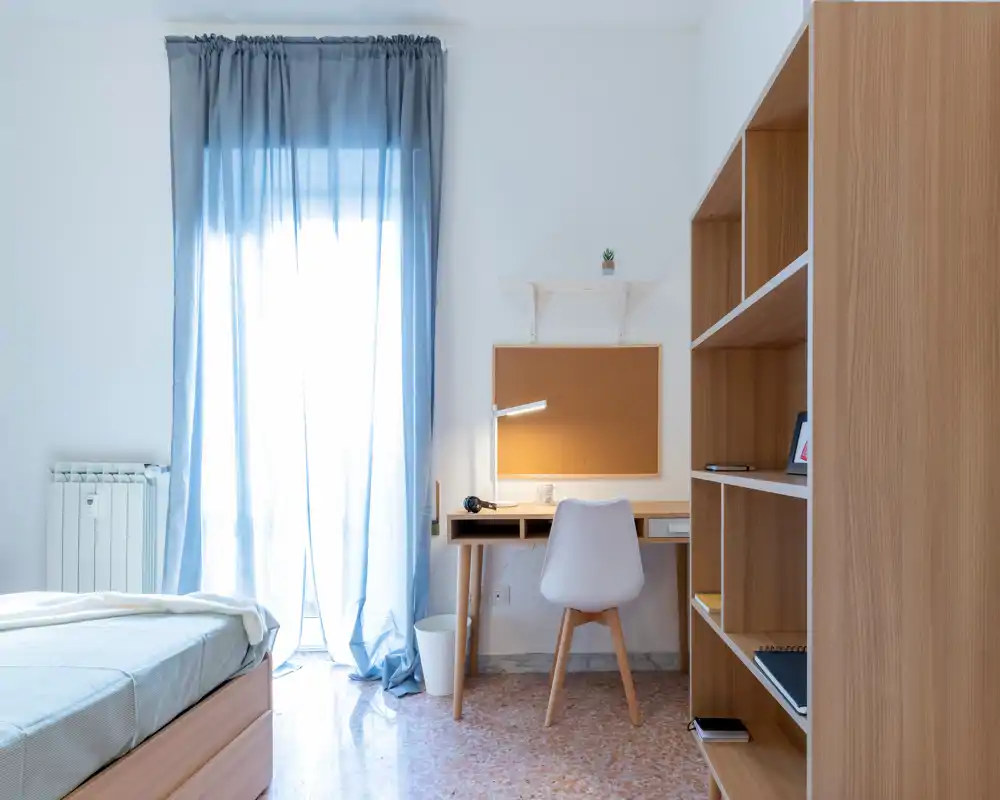 Dormitorio luminoso y ordenado con cama individual, escritorio y estanterías — ideal para estudiantes o alquileres de corta estancia. La luz natural filtrada por las cortinas azules genera un ambiente relajado.