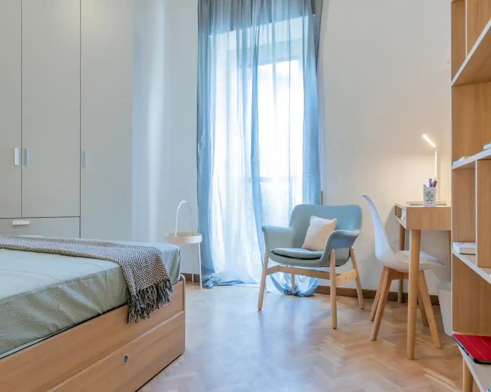 Bright, softly styled bedroom with a wooden bedframe, cozy throw and a comfortable armchair by a sheer-curtained window — ideal for restful stays.