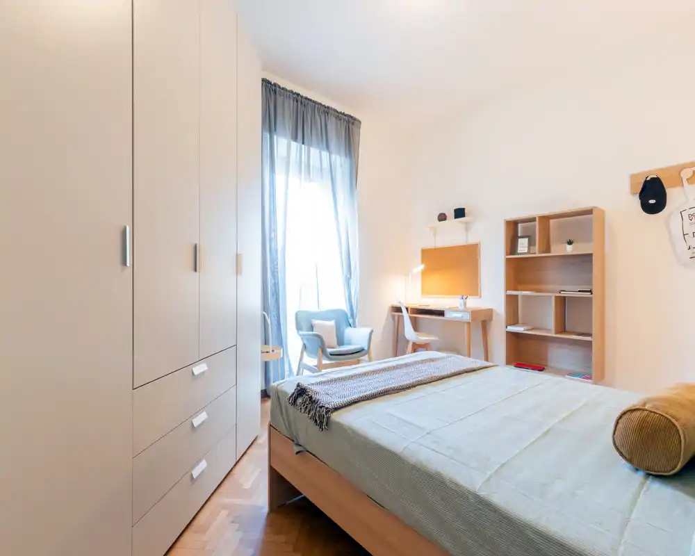Bright, well-staged bedroom with a double bed in the foreground, a comfortable armchair by the window and a functional desk and shelving—ideal for short-term rentals.