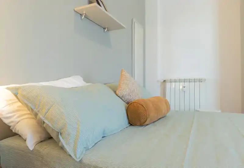 Bright, cozy bedroom view showing a well-made bed with layered pillows and soft linens in soothing pastel tones — ideal for rental listings highlighting comfort and style.