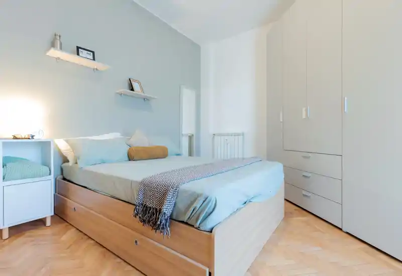 Bright, neatly staged bedroom with a wooden bed frame, pale blue linens, wardrobe and minimalist shelving; ideal for rental listings highlighting a calm, modern sleeping area.