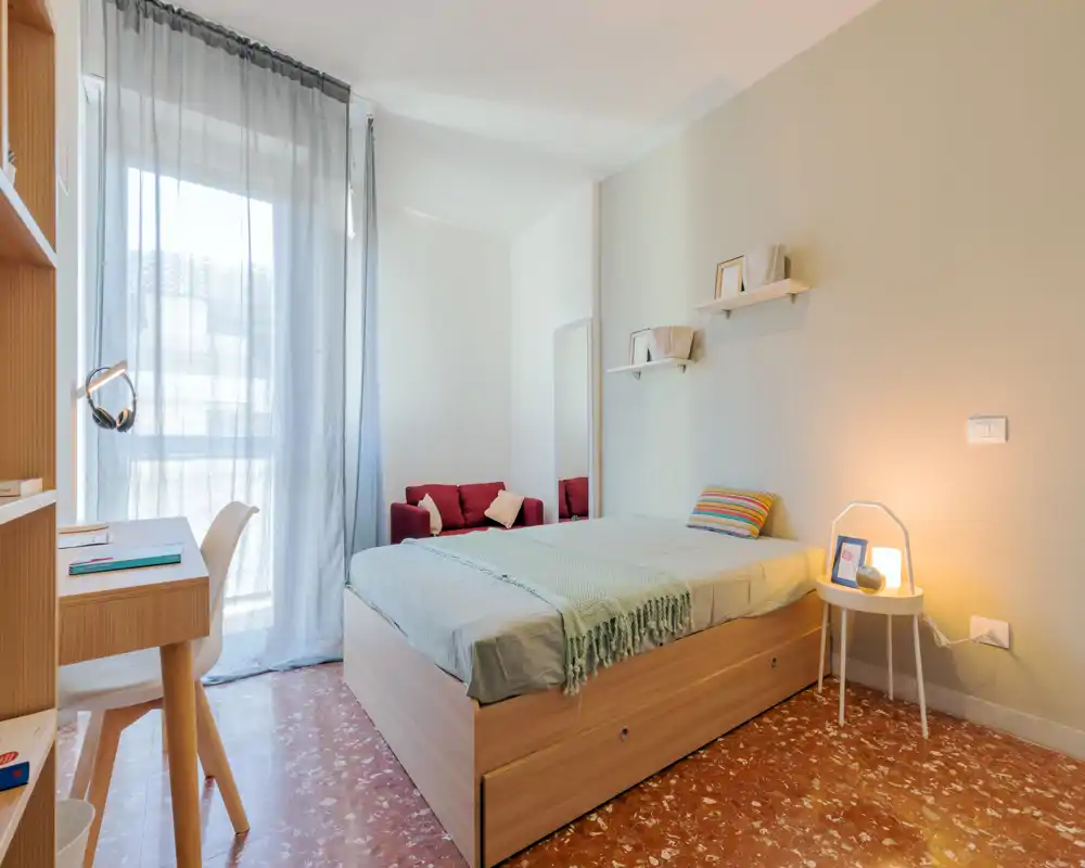 Dormitorio luminoso y moderno con cama de madera, mesita y una pequeña zona de estar junto a la ventana. La luz natural y una lámpara cálida ofrecen un ambiente acogedor.