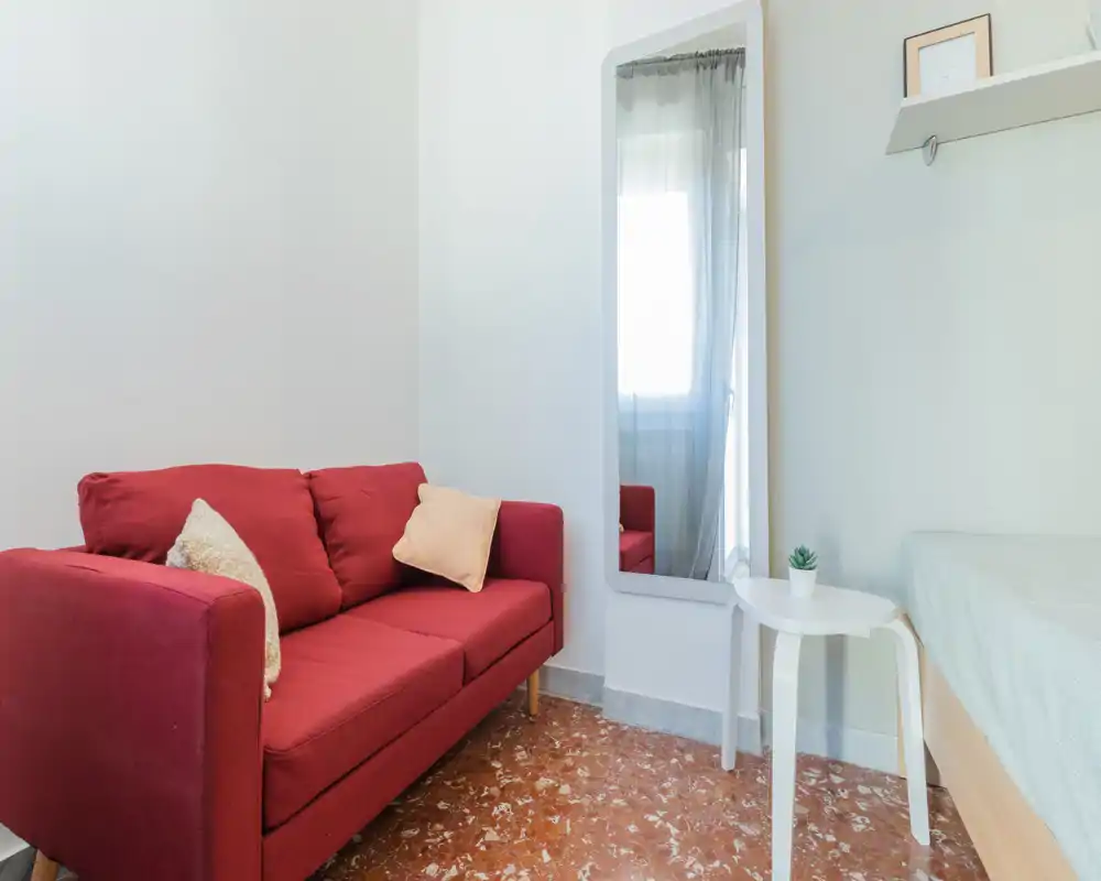 Rincón de dormitorio luminoso con sofá rojo de dos plazas, mesa auxiliar y cama parcialmente visible; decoración minimalista y luz natural.