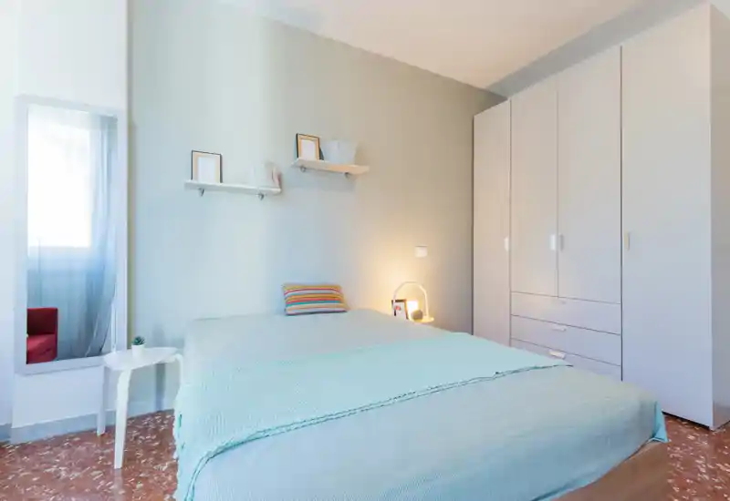 Dormitorio luminoso y de estilo sencillo con cama doble, ropa de cama en tonos pastel, lámpara de noche y gran armario — ideal para una estancia cómoda.