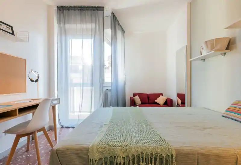 Dormitorio luminoso y bien presentado con una cama grande en primer plano, textiles suaves y un pequeño sofá rojo junto a la ventana que deja entrar abundante luz natural.