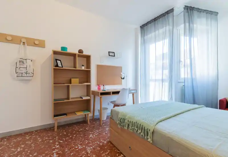 Dormitorio individual luminoso y moderno con cama de madera, manta verde claro y escritorio junto a la ventana; luz natural y estantería minimalista crean un espacio tranquilo para dormir y estudiar.