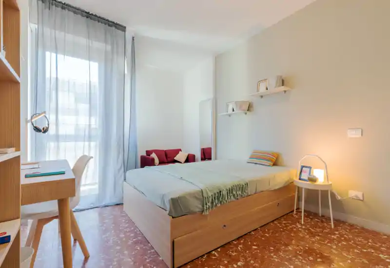 Dormitorio luminoso y moderno con cama de madera, mesita y una pequeña zona de estar junto a la ventana. La luz natural y una lámpara cálida ofrecen un ambiente acogedor.