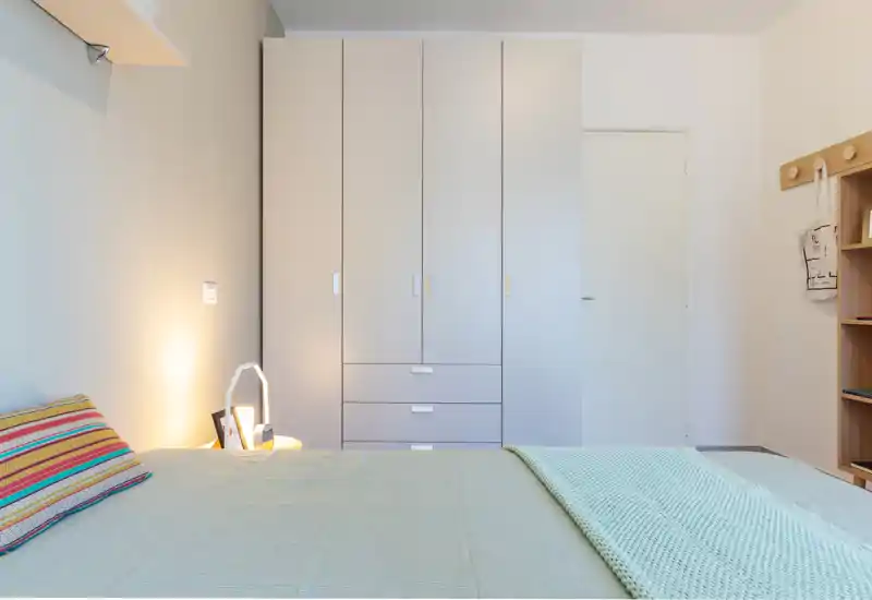 Dormitorio moderno y luminoso con cama bien hecha, textiles suaves y un amplio armario empotrado — estilo minimalista y buena iluminación ambiental.