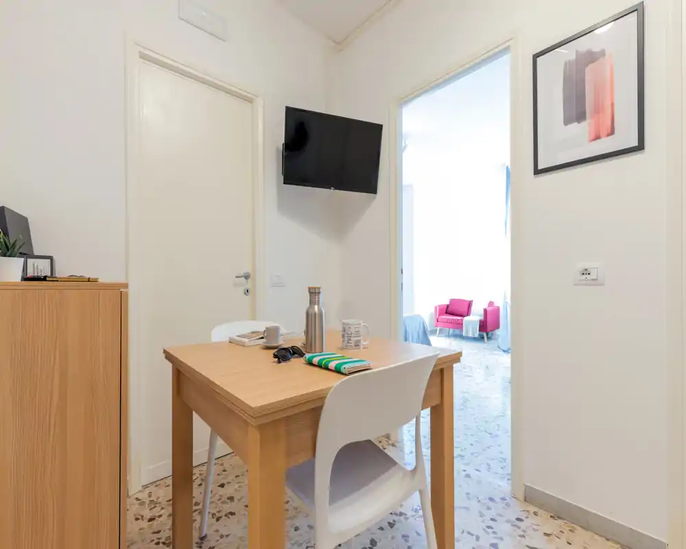 Pequeña zona de estar con una mesa de madera y dos sillas, televisor mural y una puerta que da a una habitación luminosa con un sillón rosa. Distribución limpia, moderna y práctica, ideal para estancias cortas.
