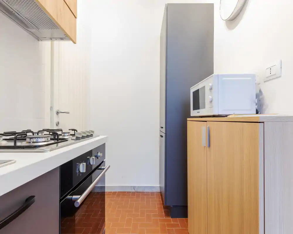 Cocina moderna y compacta con placa de gas, horno empotrado y microondas sobre un mueble de madera; paredes neutras y suelo de baldosa cálida para un espacio limpio y funcional.