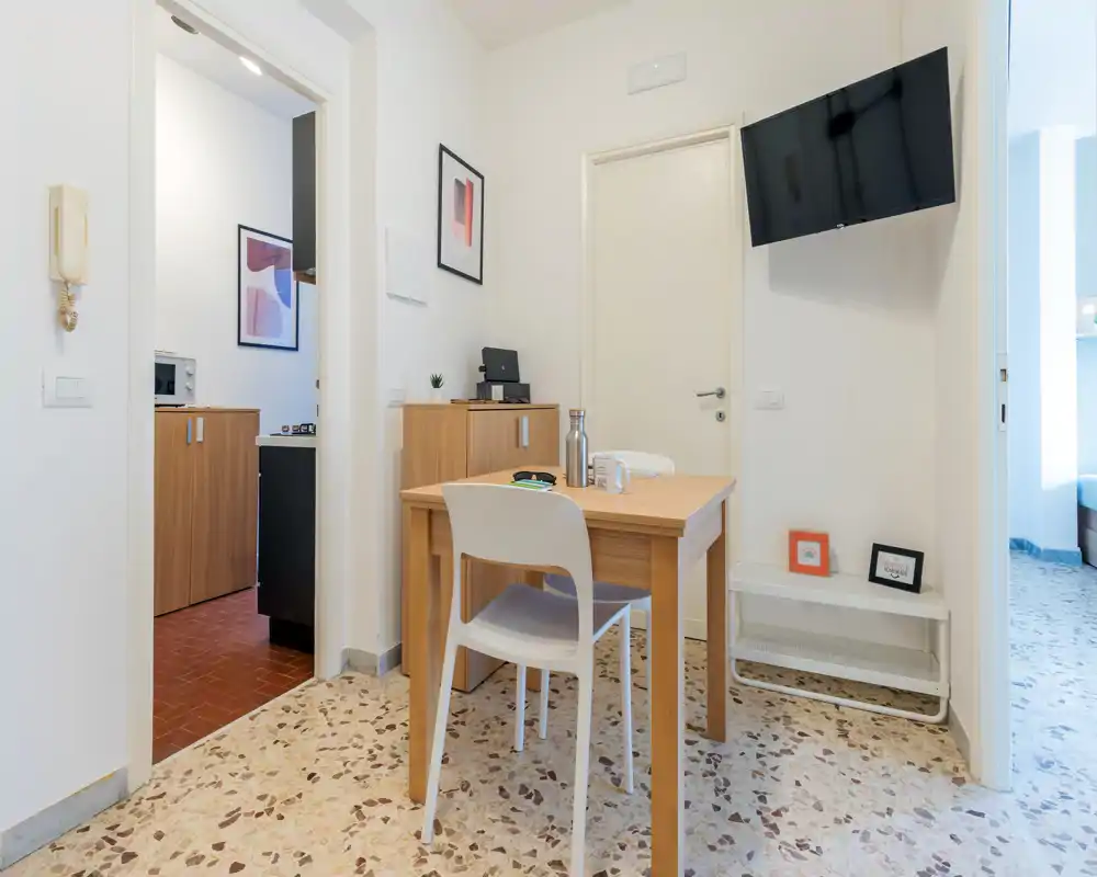 Pequeña zona de comedor con mesa de madera y dos sillas, paredes neutras y cuadros; luminosa, ordenada y adecuada para un apartamento compacto.