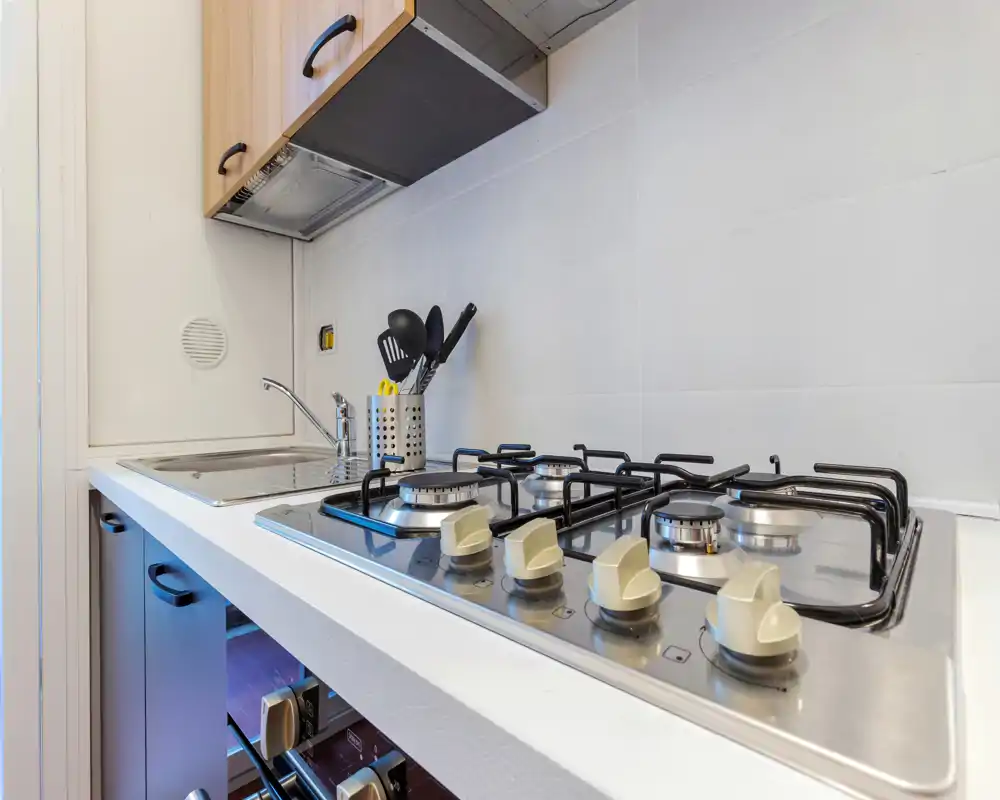Cocina compacta y moderna con encimera de gas de acero inoxidable, fregadero y soporte para utensilios; muebles de madera clara y azulejos blancos para un aspecto limpio y funcional.