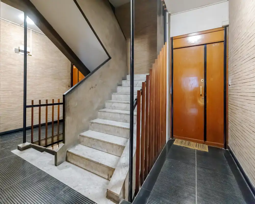 Entrada del edificio bien iluminada con escalera de mármol, puerta de madera y barandillas verticales — acceso comunitario limpio y funcional.
