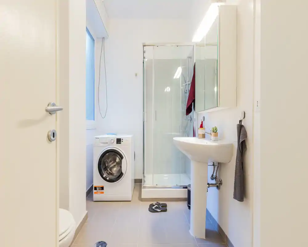 Baño moderno y luminoso con ducha de cristal, lavabo de pie y lavadora. Baldosas neutras y líneas limpias que crean un espacio funcional y fresco.