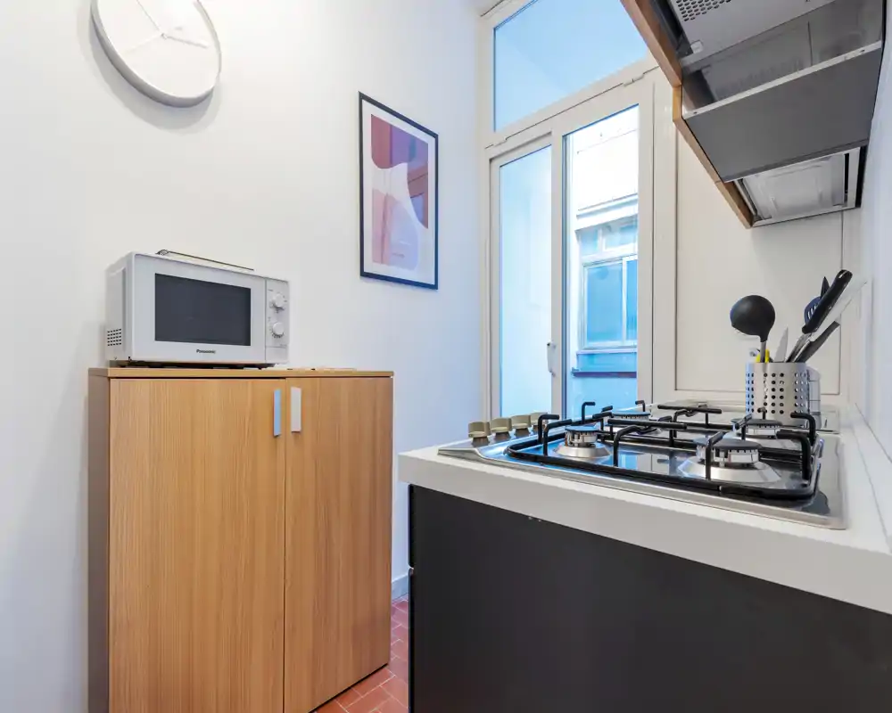 Cocina moderna y compacta con placa de gas, microondas sobre un armario de madera y una ventana luminosa que aporta luz natural—diseño eficiente para un piso pequeño.