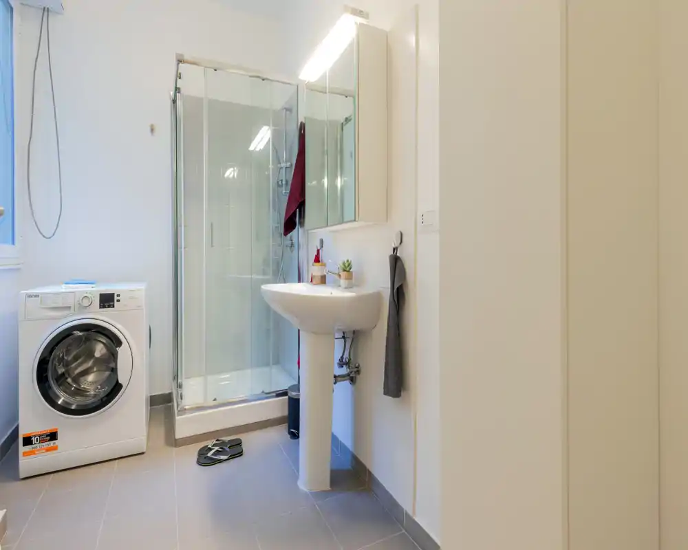 Baño moderno y luminoso con mampara de ducha de vidrio, lavabo de pie y lavadora; suelos neutros y estilo minimalista y limpio.