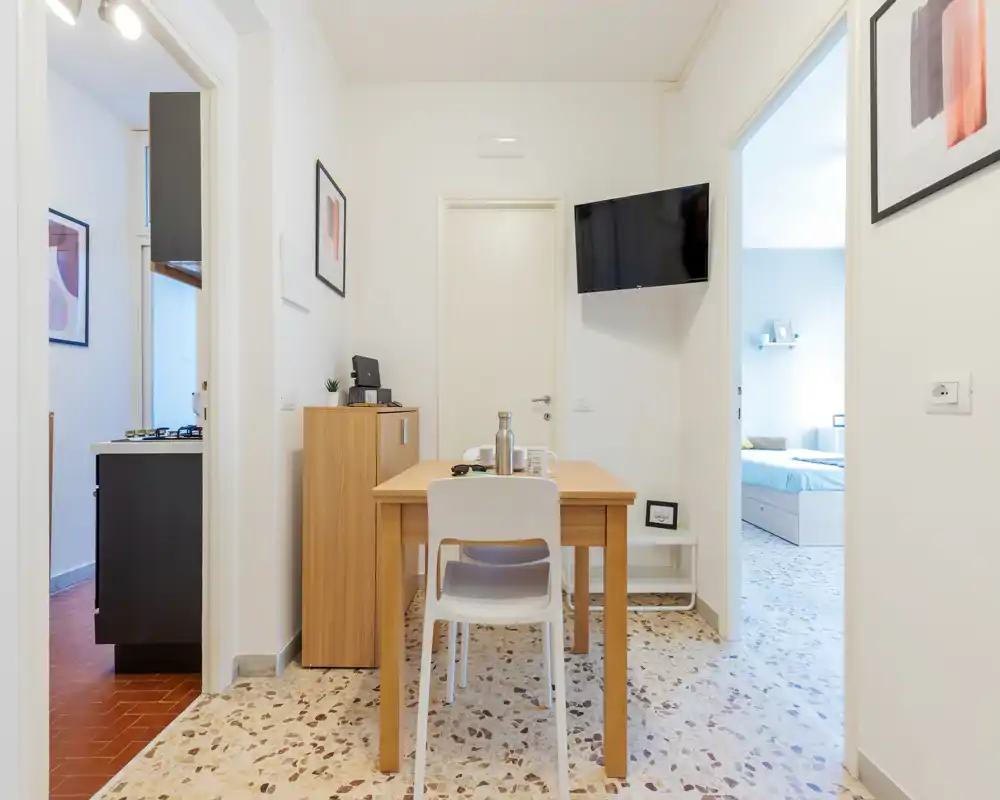 Pequeño rincón comedor/zona de estar luminosa con mesa de madera, una silla y televisor en la pared; estilo limpio y minimalista, buena luz natural.