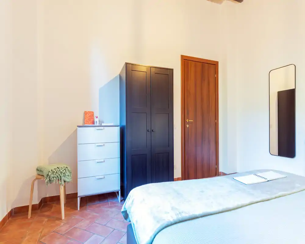 Dormitorio luminoso y de estilo sencillo con cama visible, armario y cómoda. El suelo de terracota y los textiles suaves crean un ambiente acogedor.