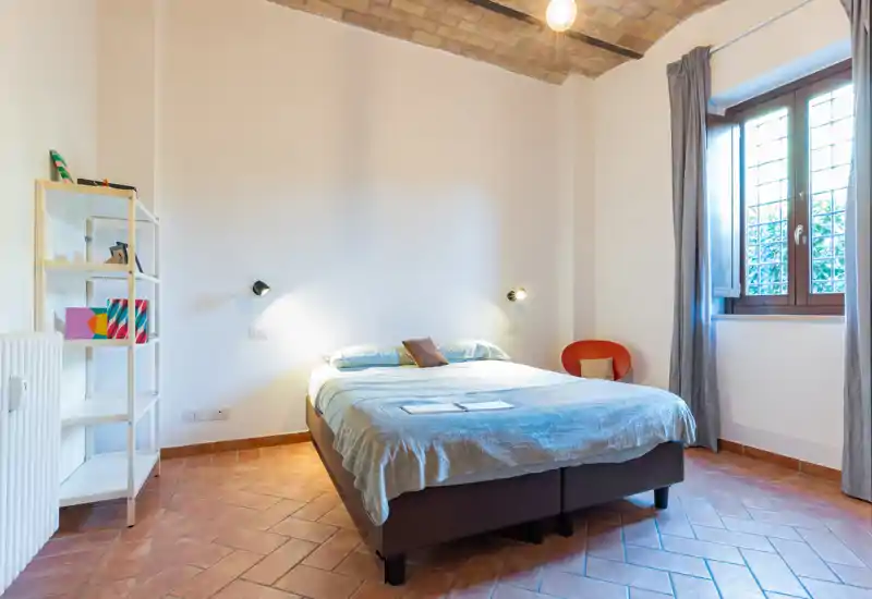 Dormitorio luminoso y bien decorado con cama doble grande, suelo de terracota y techo abovedado de ladrillo; la luz natural de la ventana y las lámparas crean un ambiente cálido y acogedor.