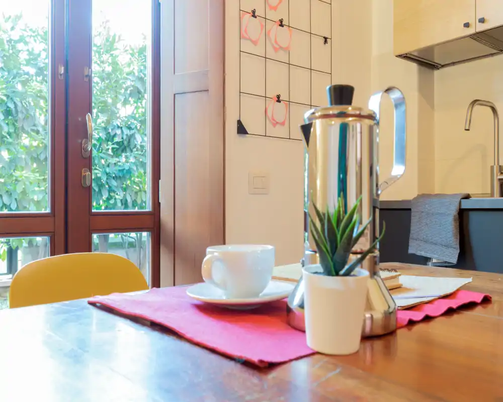 Rincón de cocina luminoso y acogedor con mesa de madera preparada para café, cafetera de acero inoxidable, taza y pequeña planta en maceta; la luz natural de la gran ventana aporta calidez.