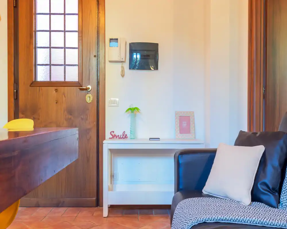 Entrada bien iluminada con puerta de madera, una pequeña consola y un sillón visible en el borde — detalles cálidos y acogedores.