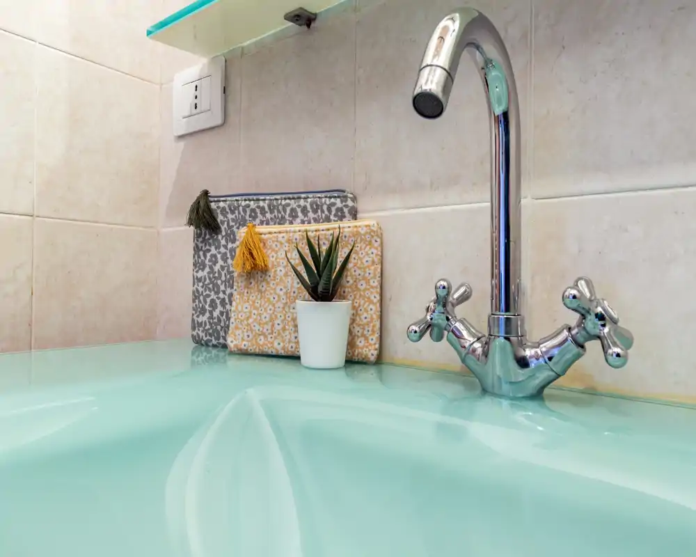 Primer plano de un lavabo de baño con grifo cromado, pequeños neceseres decorativos y una pequeña suculenta sobre una encimera color menta.