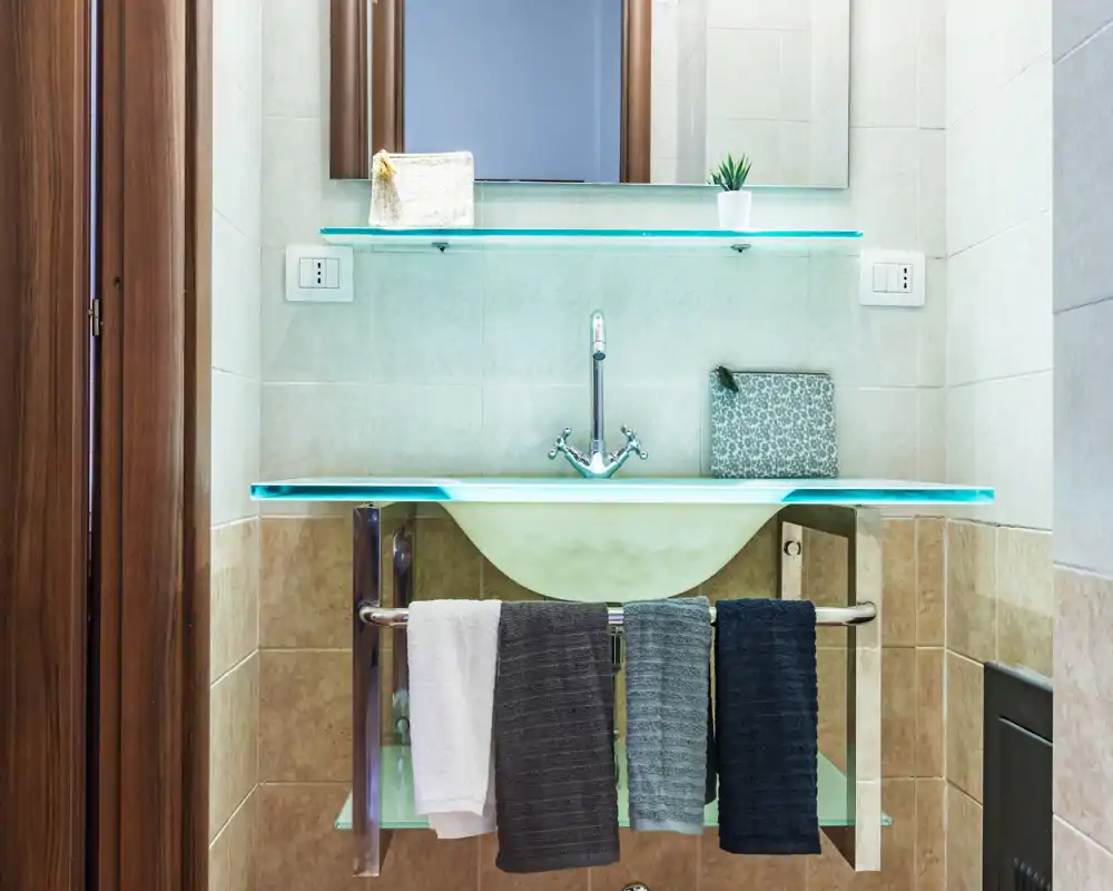 Área del lavabo de un baño moderno y luminoso con estantes de vidrio, grifo central y toallas colgadas ordenadamente; azulejos limpios y detalles decorativos sencillos.