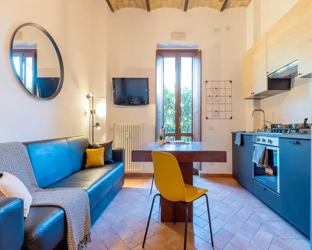 Área de estar luminosa y con estilo con un cómodo sofá, televisor en la pared y cocina integrada. Suelo de terracota, mesa de madera y luz natural desde la ventana alta, ideal para un apartamento acogedor.