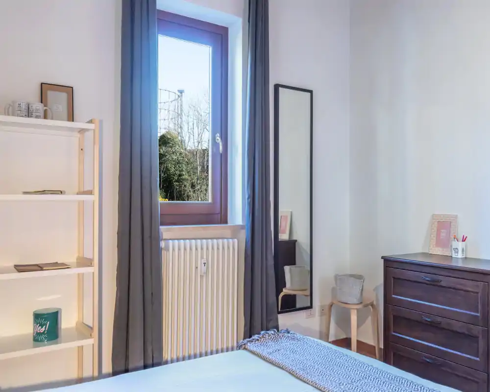 Chambre lumineuse au style épuré avec lumière naturelle par la fenêtre, lit visible avec plaid texturé, commode en bois et miroir en pied.