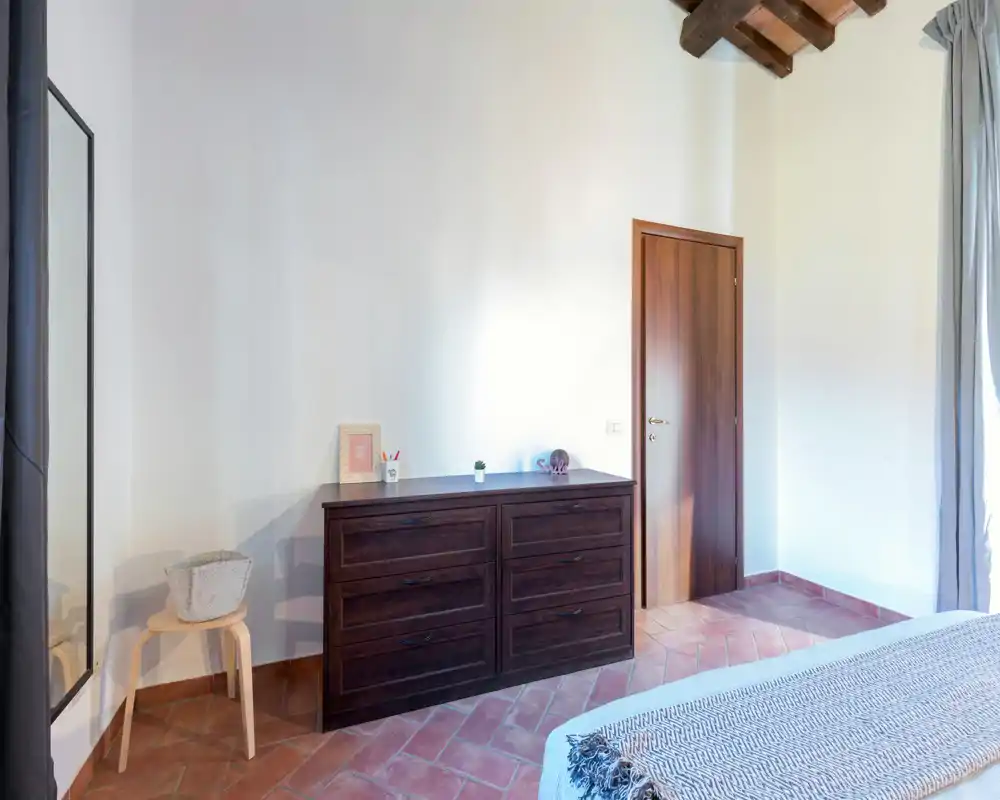 Coin chambre lumineux et chaleureux avec commode en bois, sol en terre cuite et lit partiellement visible avec textiles doux.
