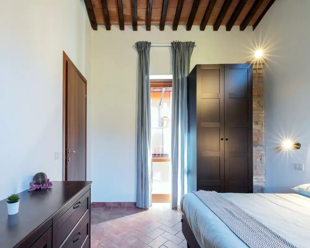Chambre lumineuse et bien composée avec poutres apparentes, lit double partiellement visible à droite, armoire et commode en bois foncé, et une grande fenêtre avec rideaux laissant entrer la lumière naturelle.