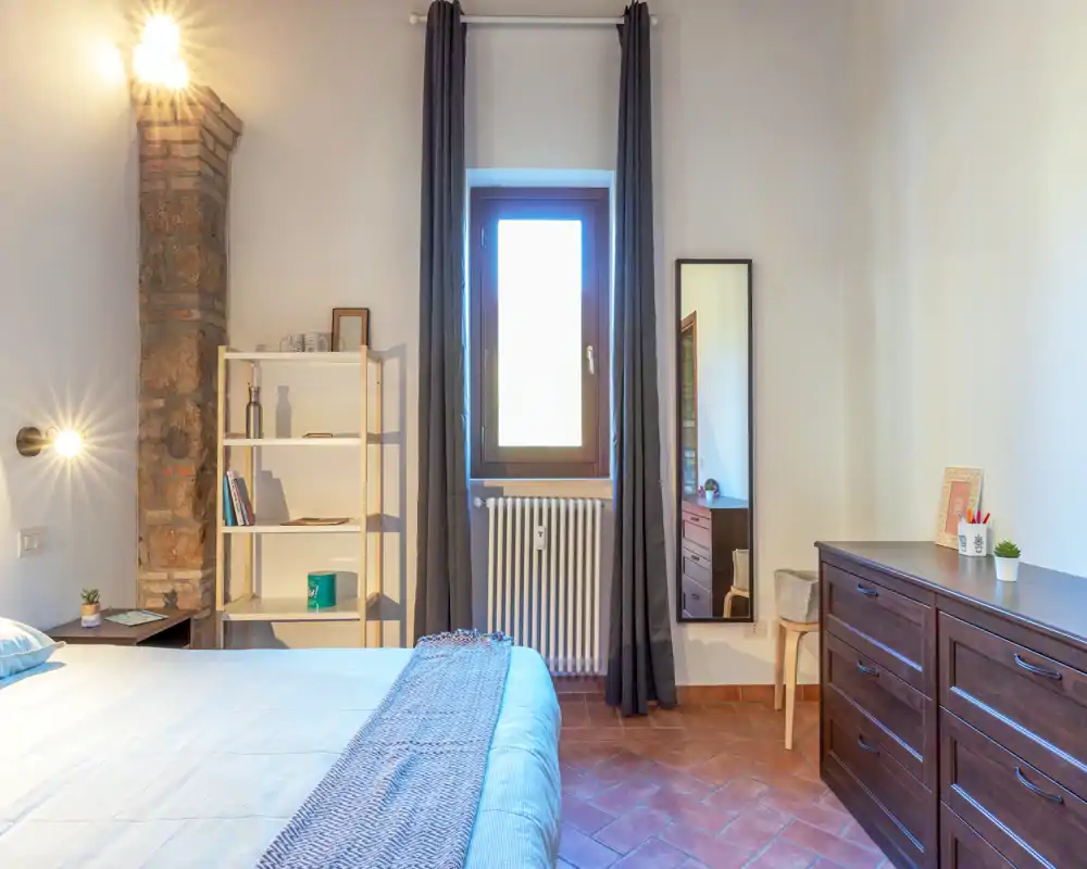 Chambre lumineuse et chaleureuse avec grand lit, étagère près du chevet, miroir vertical et commode en bois. La lumière naturelle et les lampes créent une atmosphère accueillante.