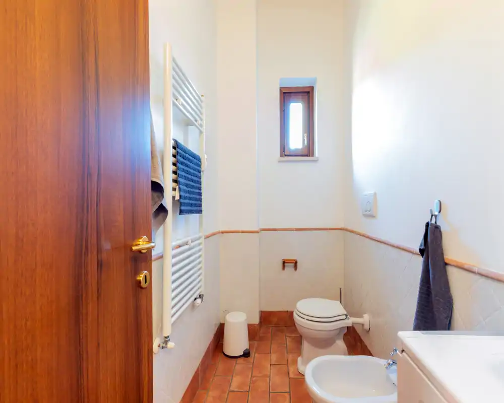 Salle de bains lumineuse avec WC, bidet et sol carrelé ; petite fenêtre pour la lumière naturelle et un radiateur sèche-serviettes mural.