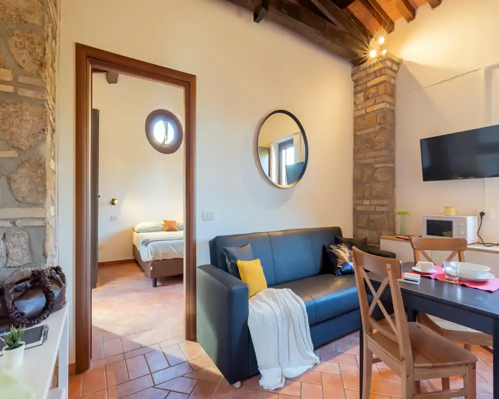 Pièce de vie lumineuse et bien aménagée avec un canapé foncé, coussins décoratifs et une table à manger; une porte ouverte laisse voir une chambre cosy. Les poutres apparentes et la pierre apportent du caractère rustique.